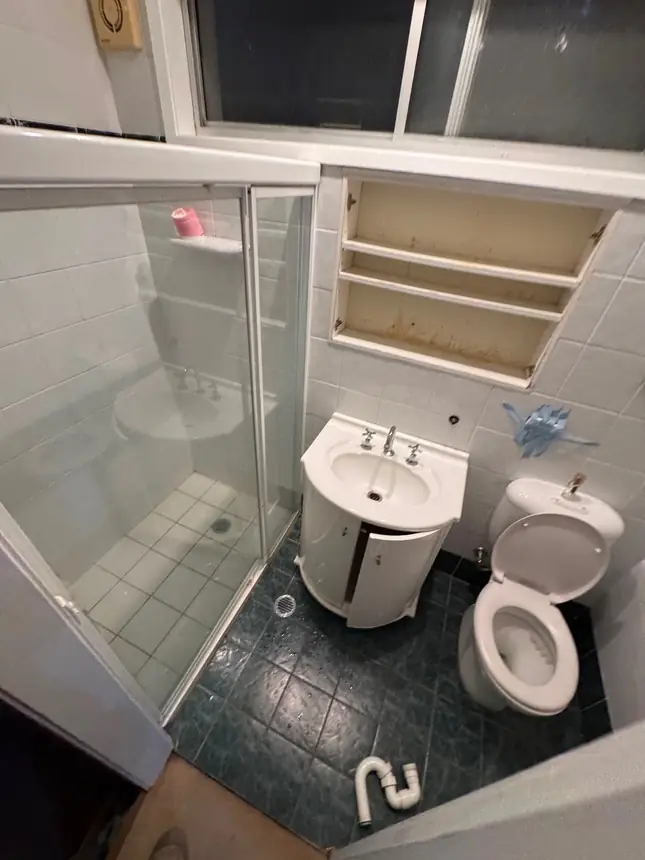 A small dated bathroom before renovation, with dark floor tiles and a corner shower